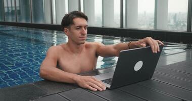 Freelancer man is working at a laptop as digital nomad while traveling. Male freelancer opens laptop while resting at rooftop swimming pool at skyscraper. Business male working on computer on vacation video